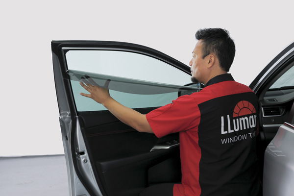 A worker with a Llumar shirt installs a window film on a car’s window Caption: Source - Innovative Film Werks