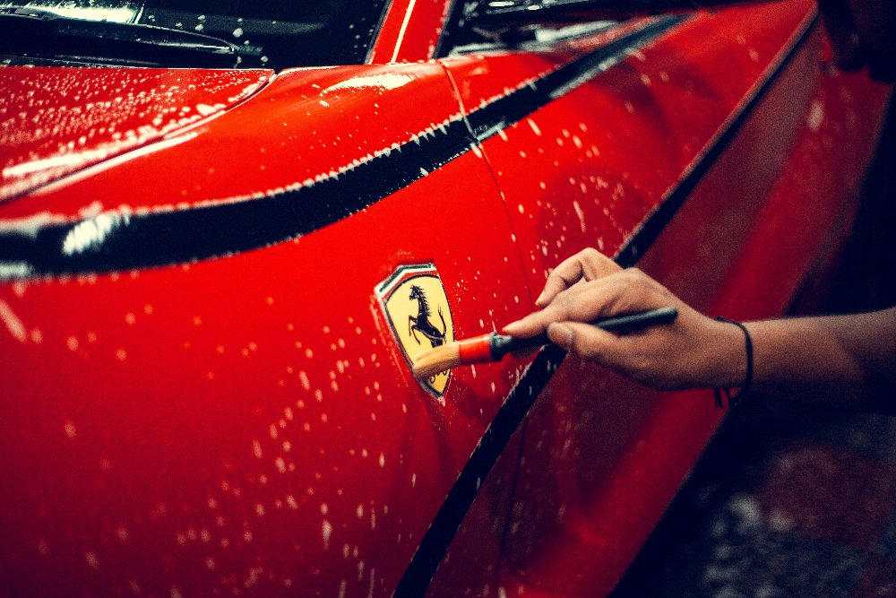 A car detailer works on the logo of a red Ferrari with a detailing brush