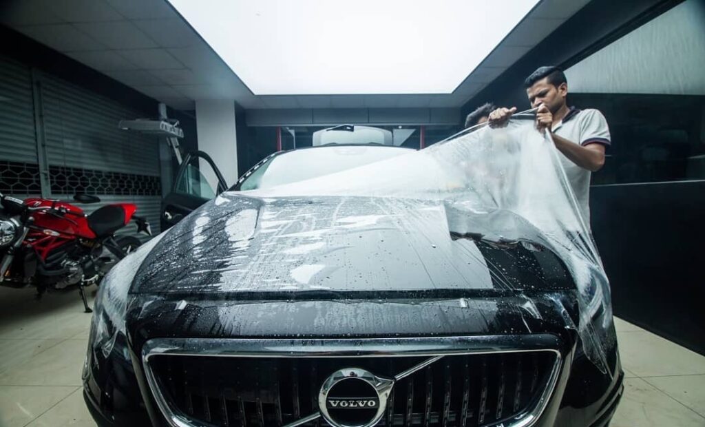 A worker applies clear PPF on the bonnet of a black coloured Volvo sedan