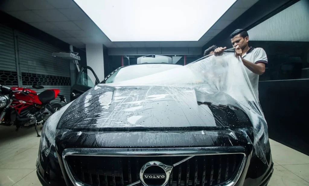 A worker applies car PPF on the back panel of the rear view mirror of a car inside a detailing studio
