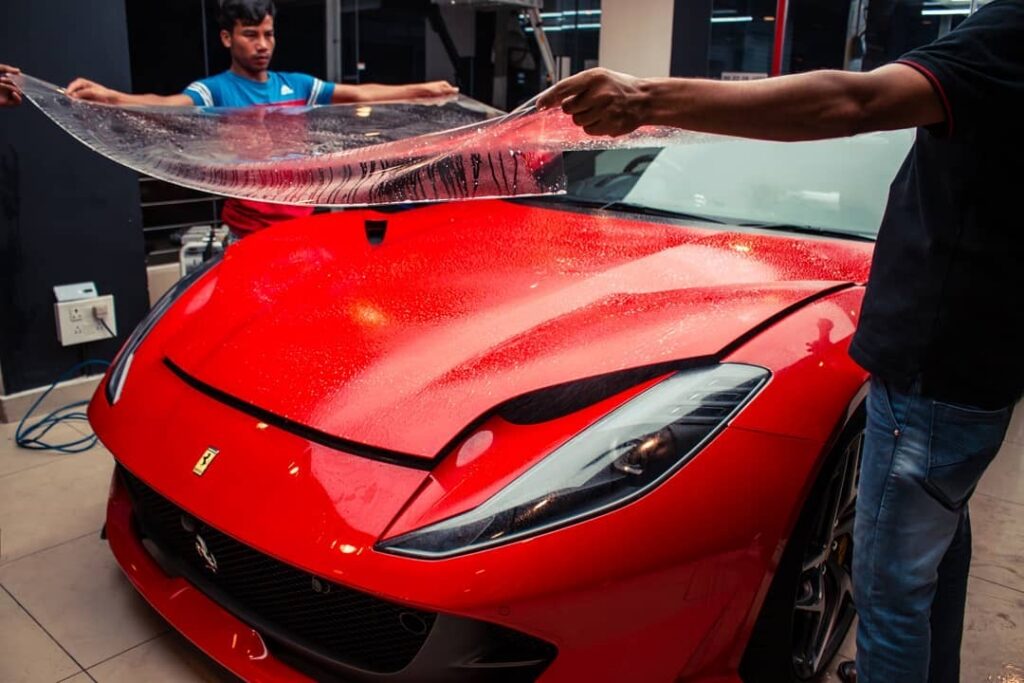 Two workers place a clear PPF on the bonnet of a Ferrari sedan