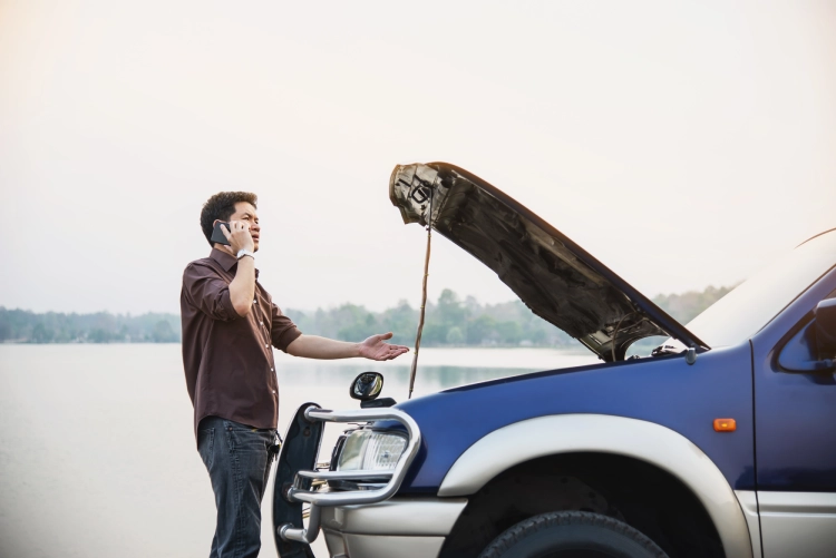 Man standing beside a car with the bonnet open, talking on the phone near a lakeside road.