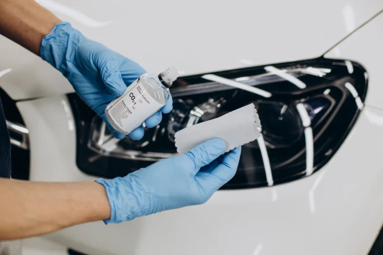 Close-up shot of a worker holding up a bottle of ceramic coating and an applicator, with a white sedan in the background.