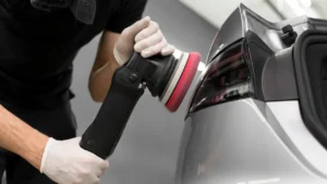 A car detailer performing paint correction on a silver car's back panel under professional studio lighting at a car detailing studio