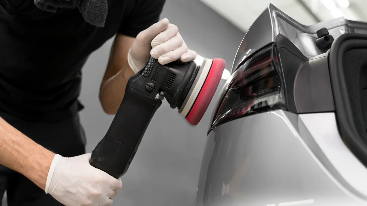A car detailer performing paint correction on a silver car's back panel under professional studio lighting at a car detailing studio